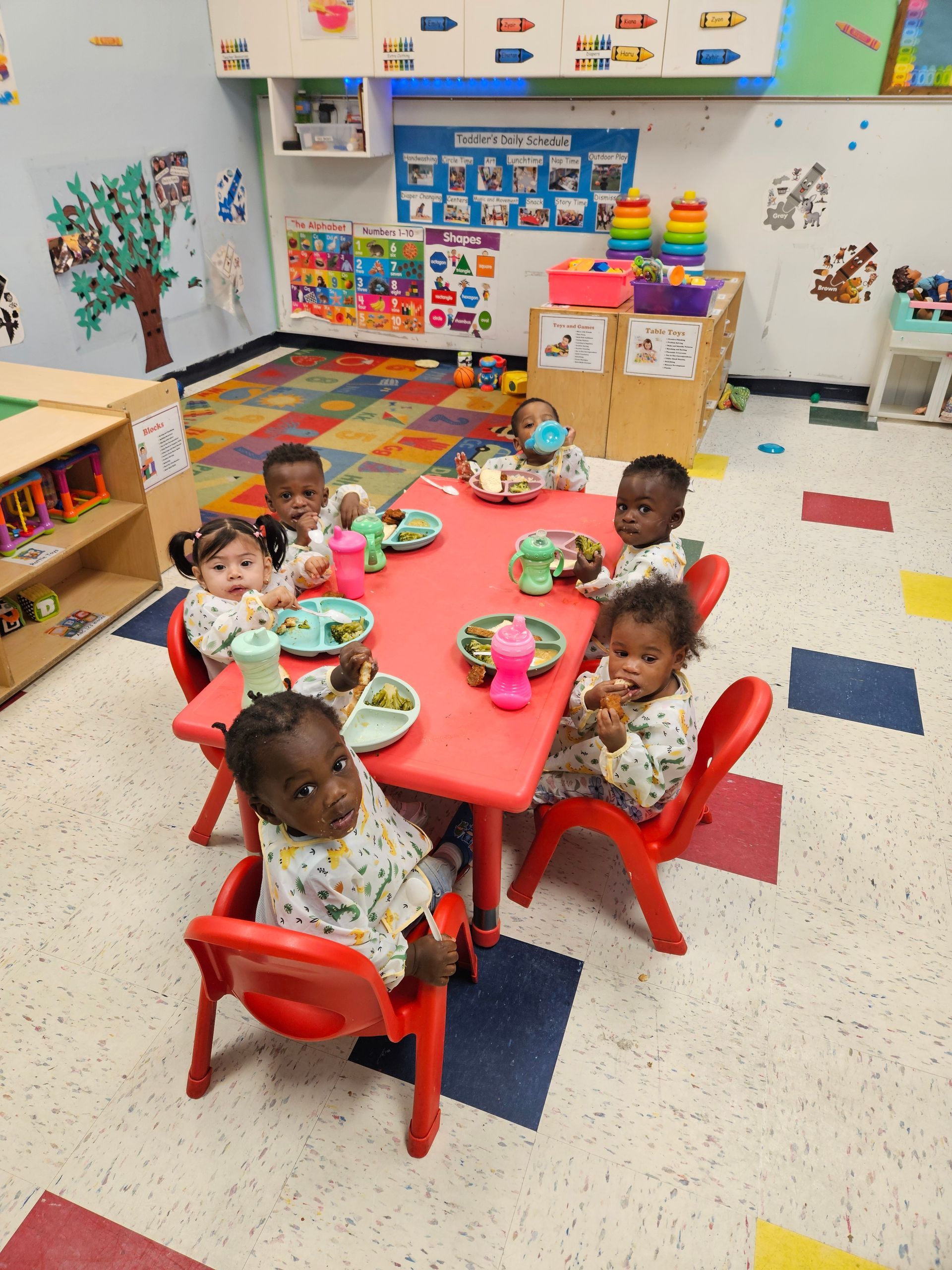 Six babies in pajamas sit at a red table, eating in a colorful classroom.