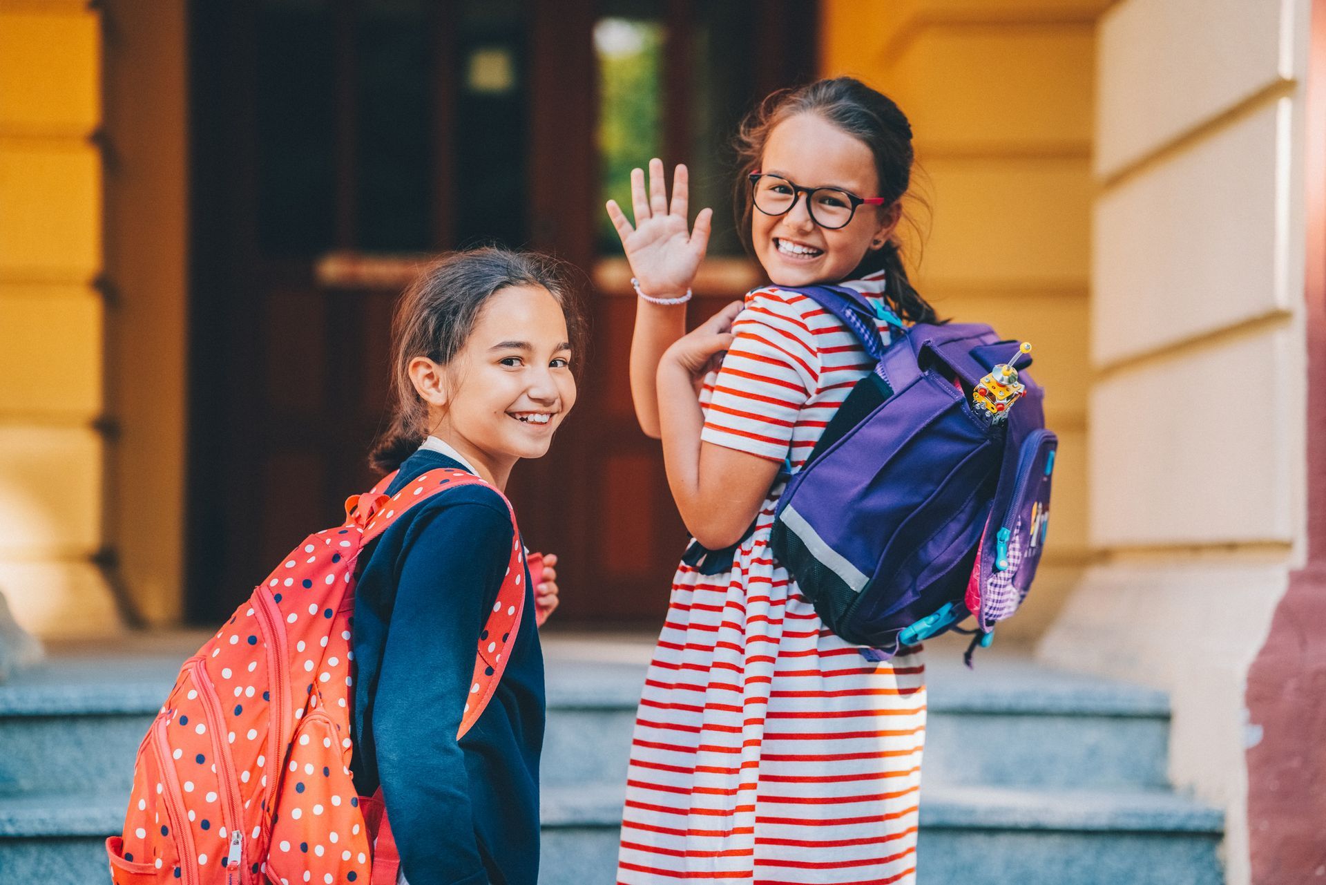 Two girls with backpacks wave in front of a school building.