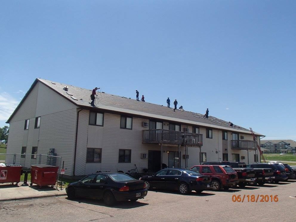 Roofers working on a two-story apartment building, sunny day. Cars parked in front.