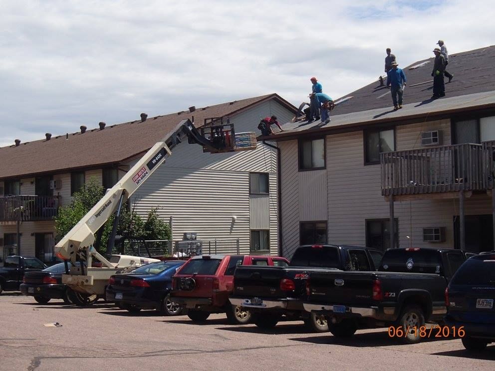Construction workers on a roof, using a lift, repairing an apartment building on a sunny day. Cars parked below.