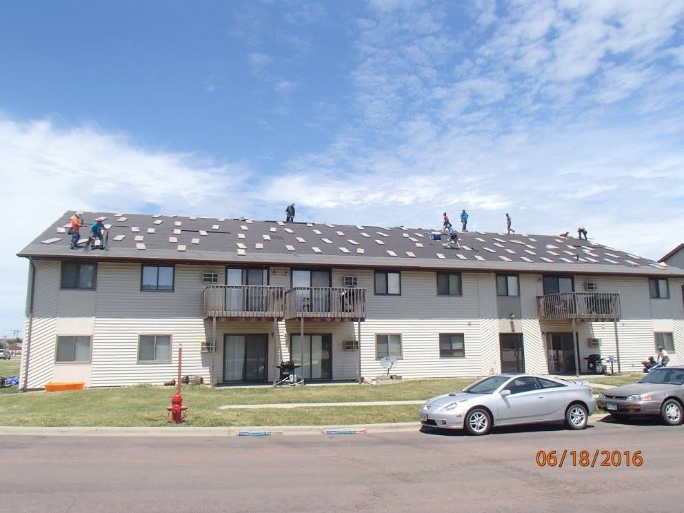 Roofers on a two-story building with partially removed roof shingles; parked cars in front. Blue sky.