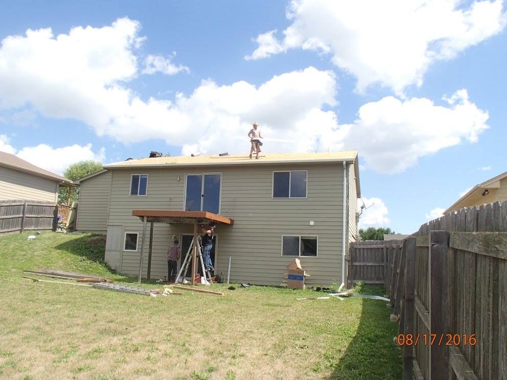Workers on roof of a two-story house, with a deck below and a fenced backyard, under a cloudy sky.