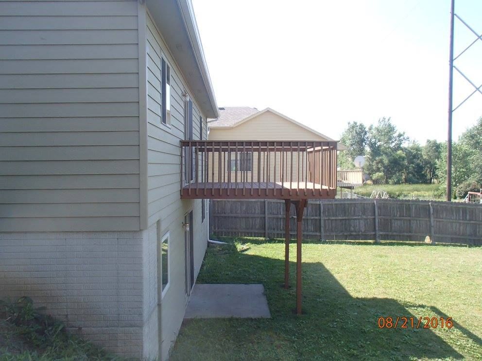 A wooden deck attached to a tan house, supported by posts, overlooking a grassy yard and a wooden fence.