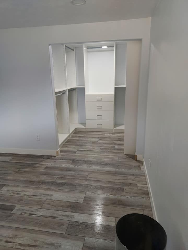 Walk-in closet with white shelves and drawers, accessible from a room with gray wood-look flooring.
