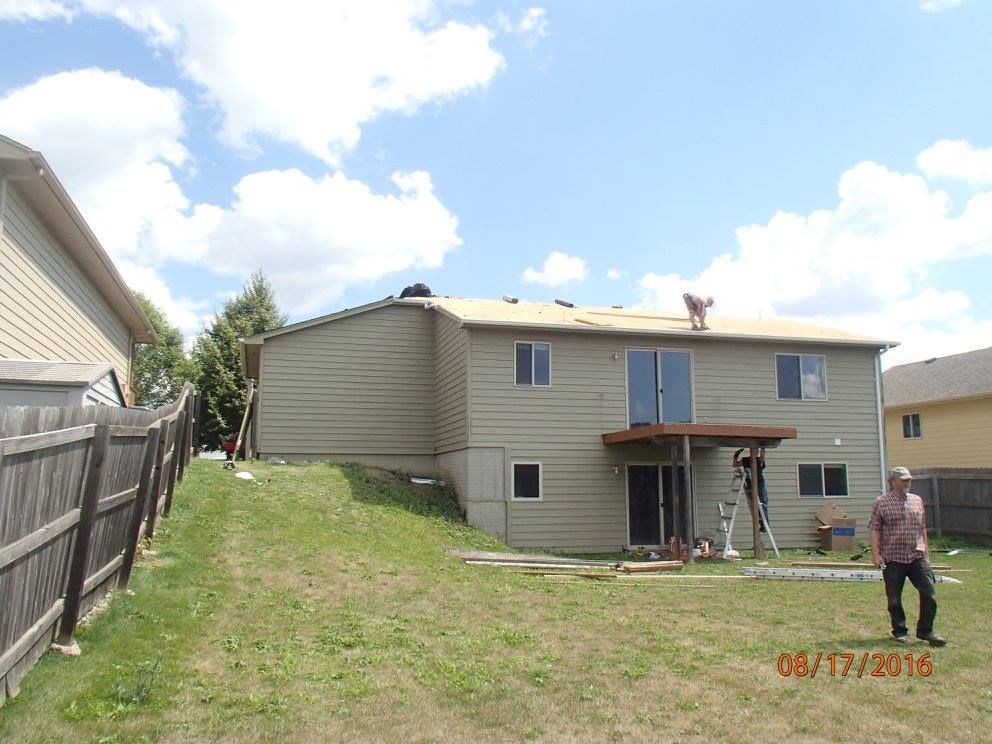 Backyard view of a two-story house with roof replacement in progress on a sunny day. A person stands in the yard.
