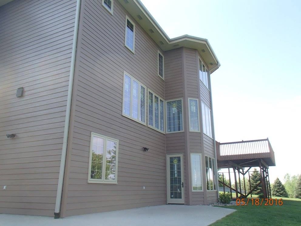 Brown siding house with many windows, a small deck, and a concrete patio on a sunny day.