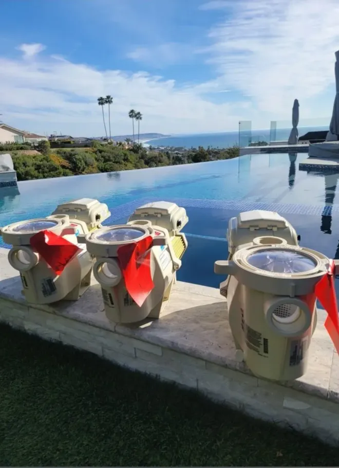Three beige pool lights with red ribbons sit on a pool ledge overlooking the ocean and palm trees.