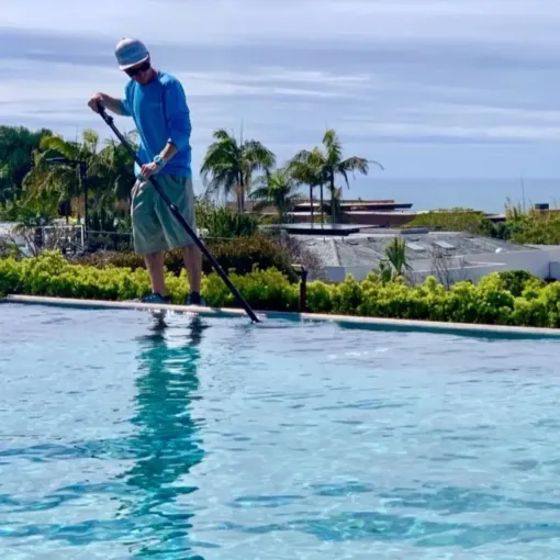 Man cleaning pool with long-handled net, blue shirt, tan shorts, sunny outdoor setting.