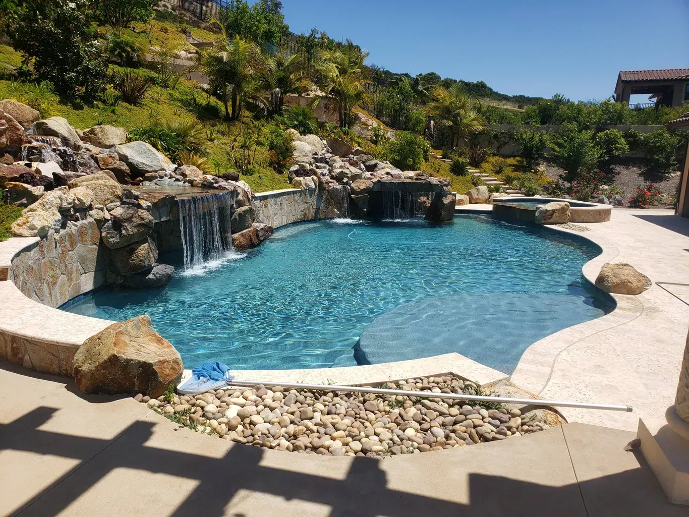 Swimming pool with waterfalls, rocky accents, and a hillside backdrop on a sunny day.