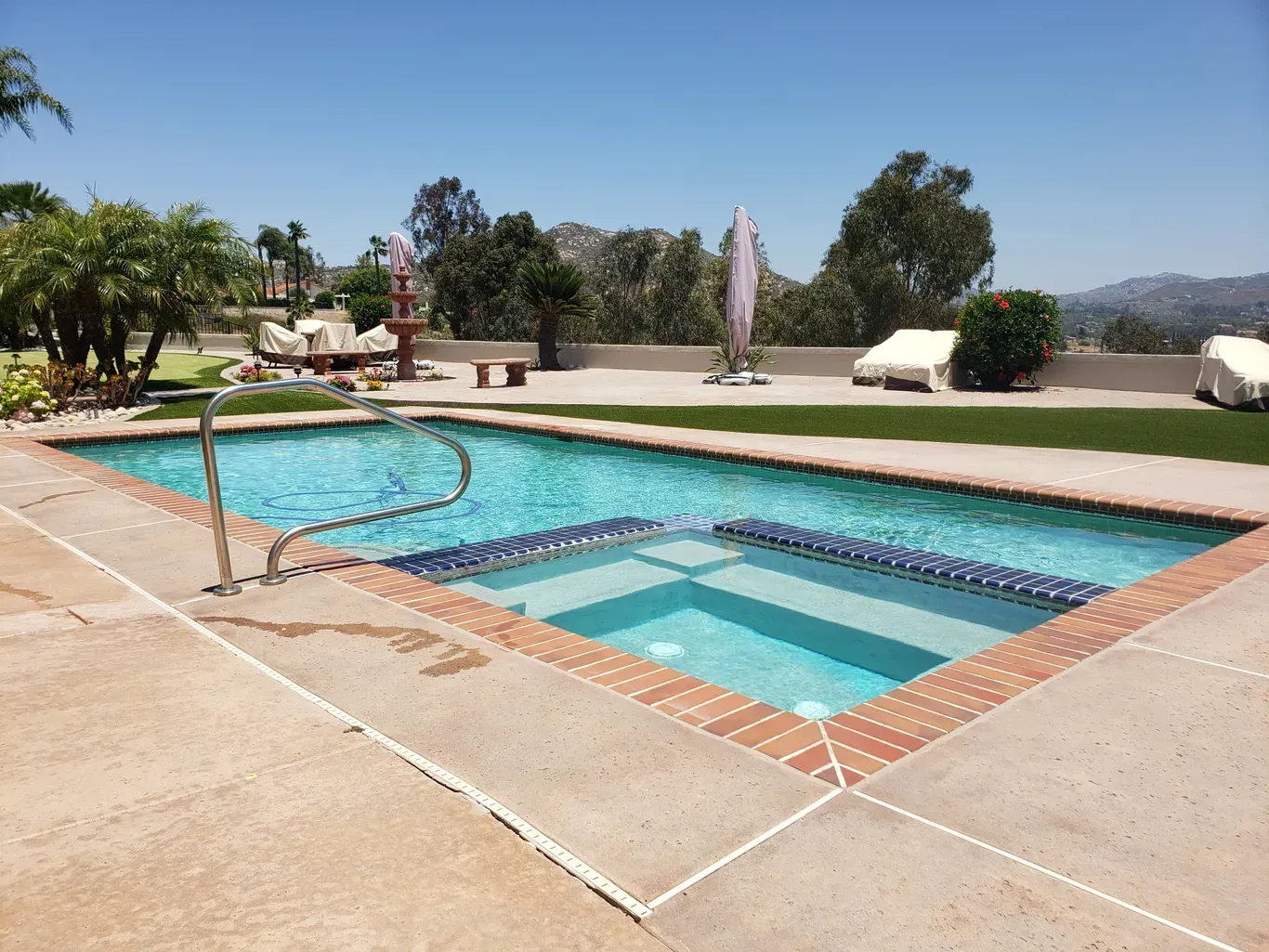 Swimming pool with jacuzzi, surrounded by concrete, grass, and trees, mountains in background.