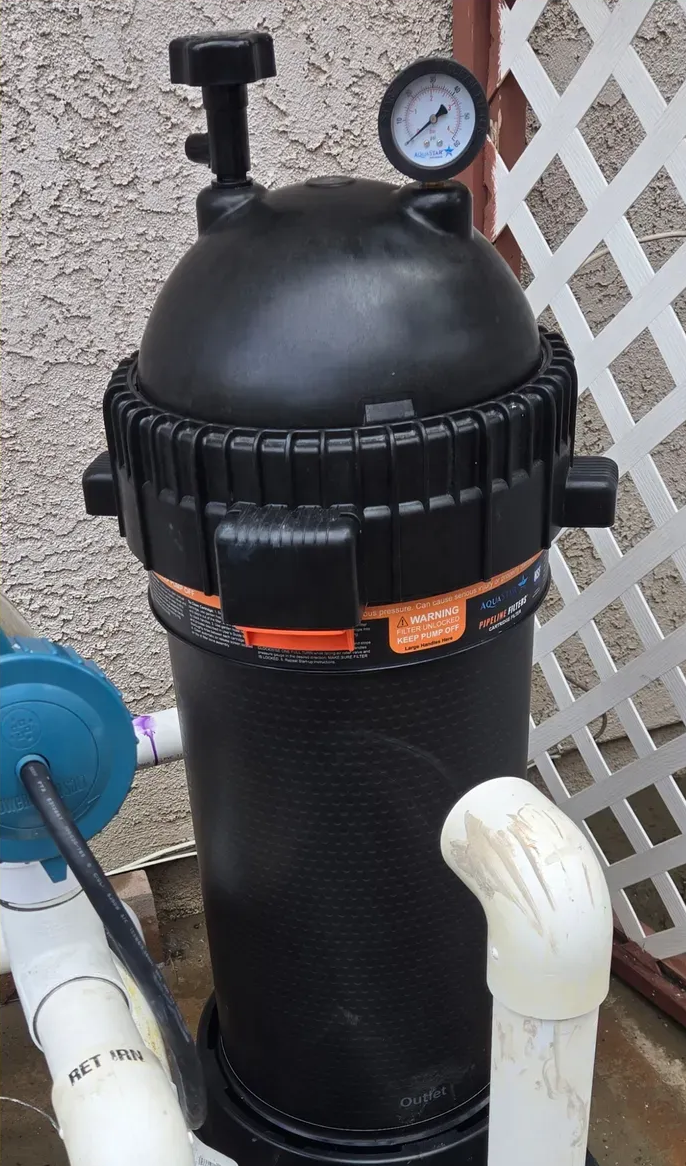 Black pool filter with pressure gauge, attached to white and blue pipes, outdoor setting.