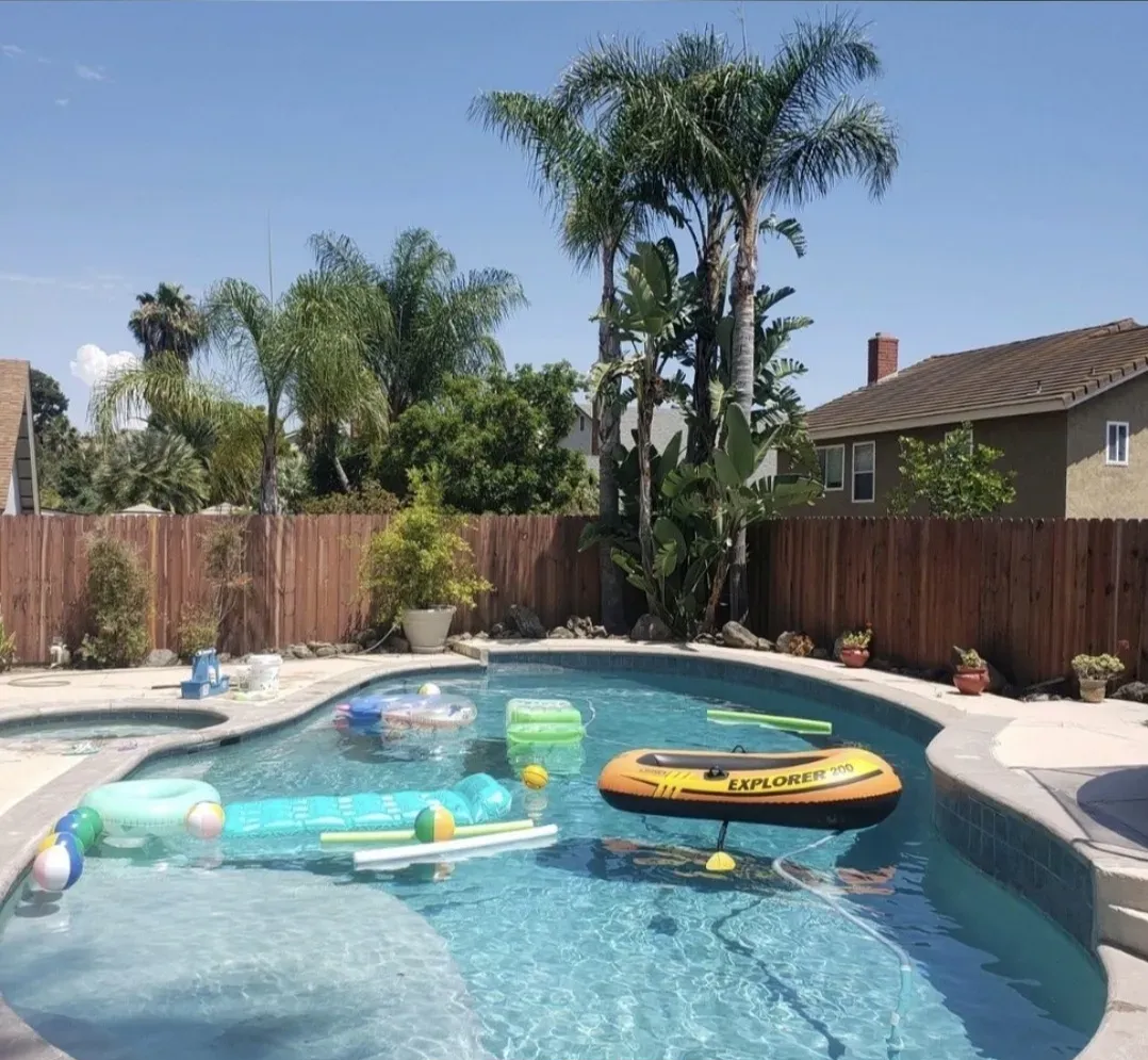 Pool with floaties and an inflatable boat in a sunny backyard, surrounded by a wooden fence and palm trees.