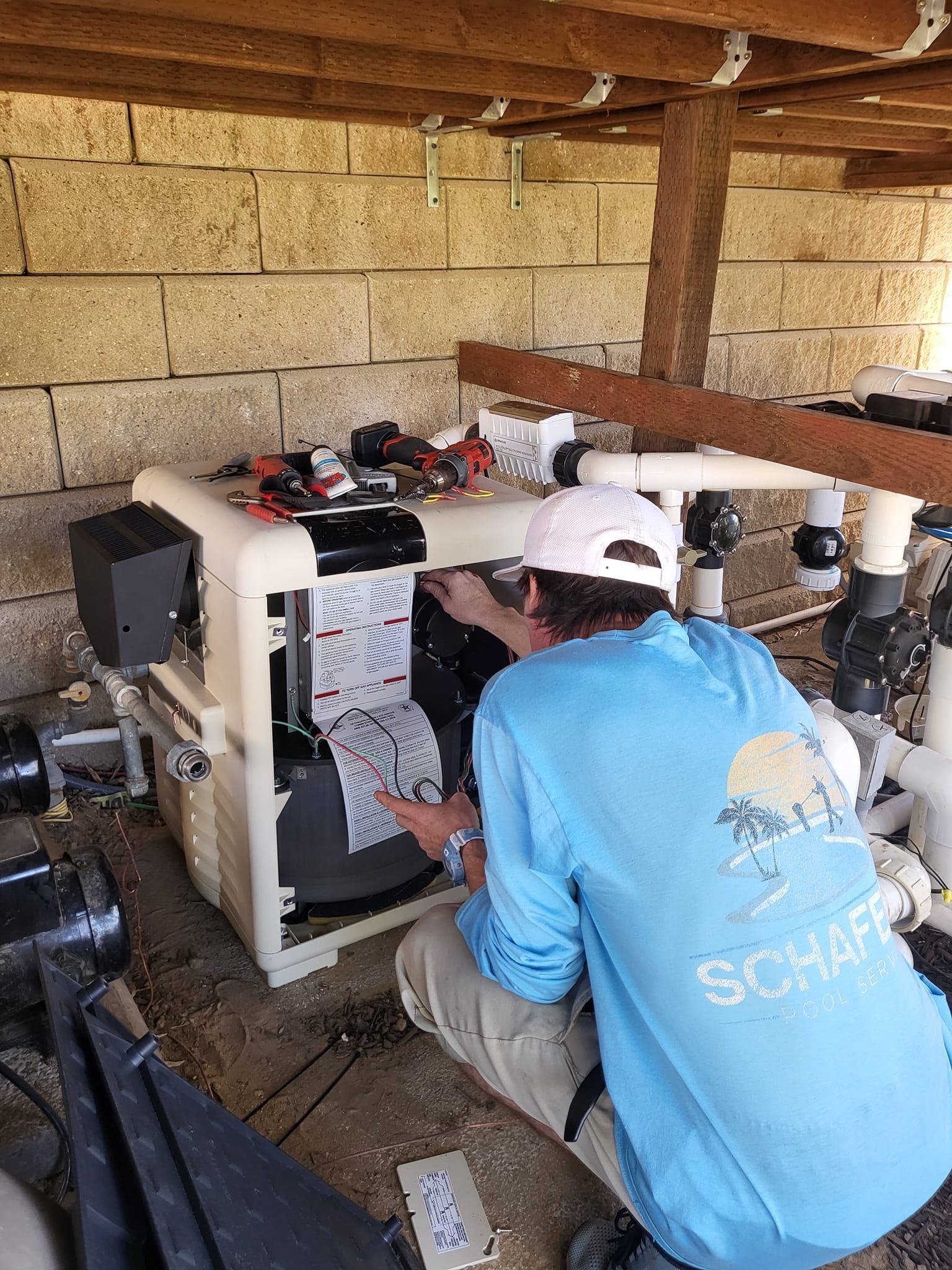 Person in blue shirt fixing pool heater under a wooden structure. Beige brick wall.