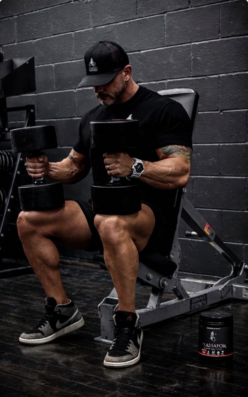 A person wearing a black t-shirt and cap sits on a gym bench, resting heavy dumbbells on their thighs.