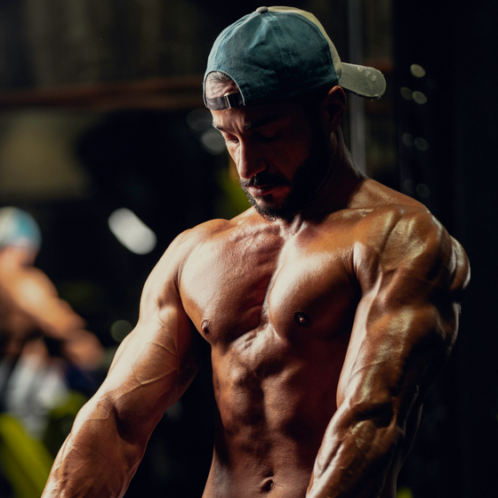 Muscular man in a backward cap posing in a dim gym, lit with dramatic shadows