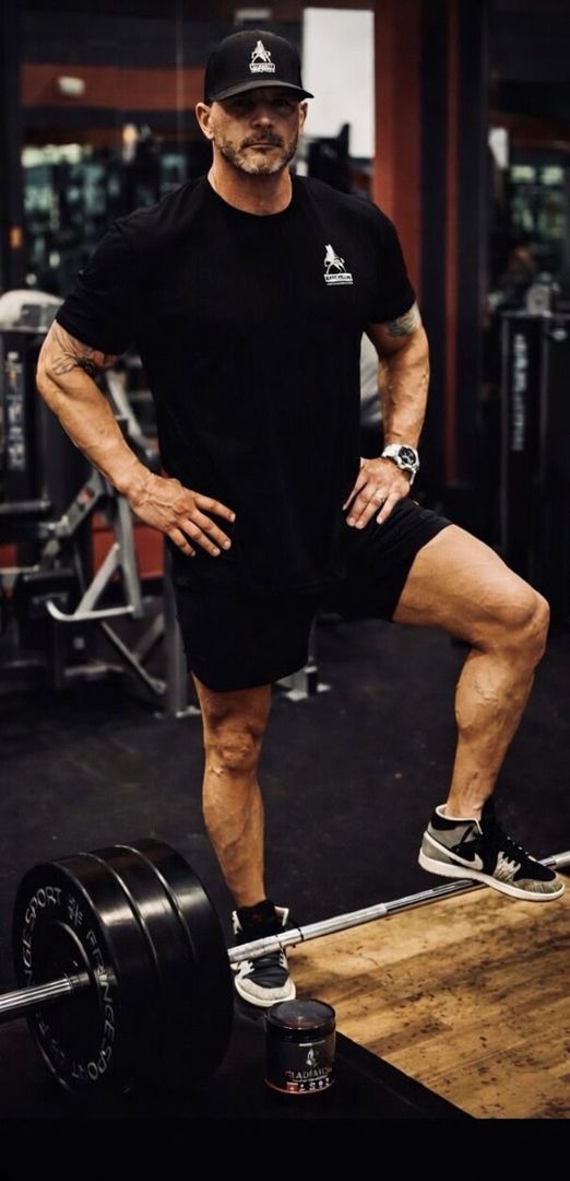 A person in a black shirt and cap posing in a gym with one leg resting on a barbell.