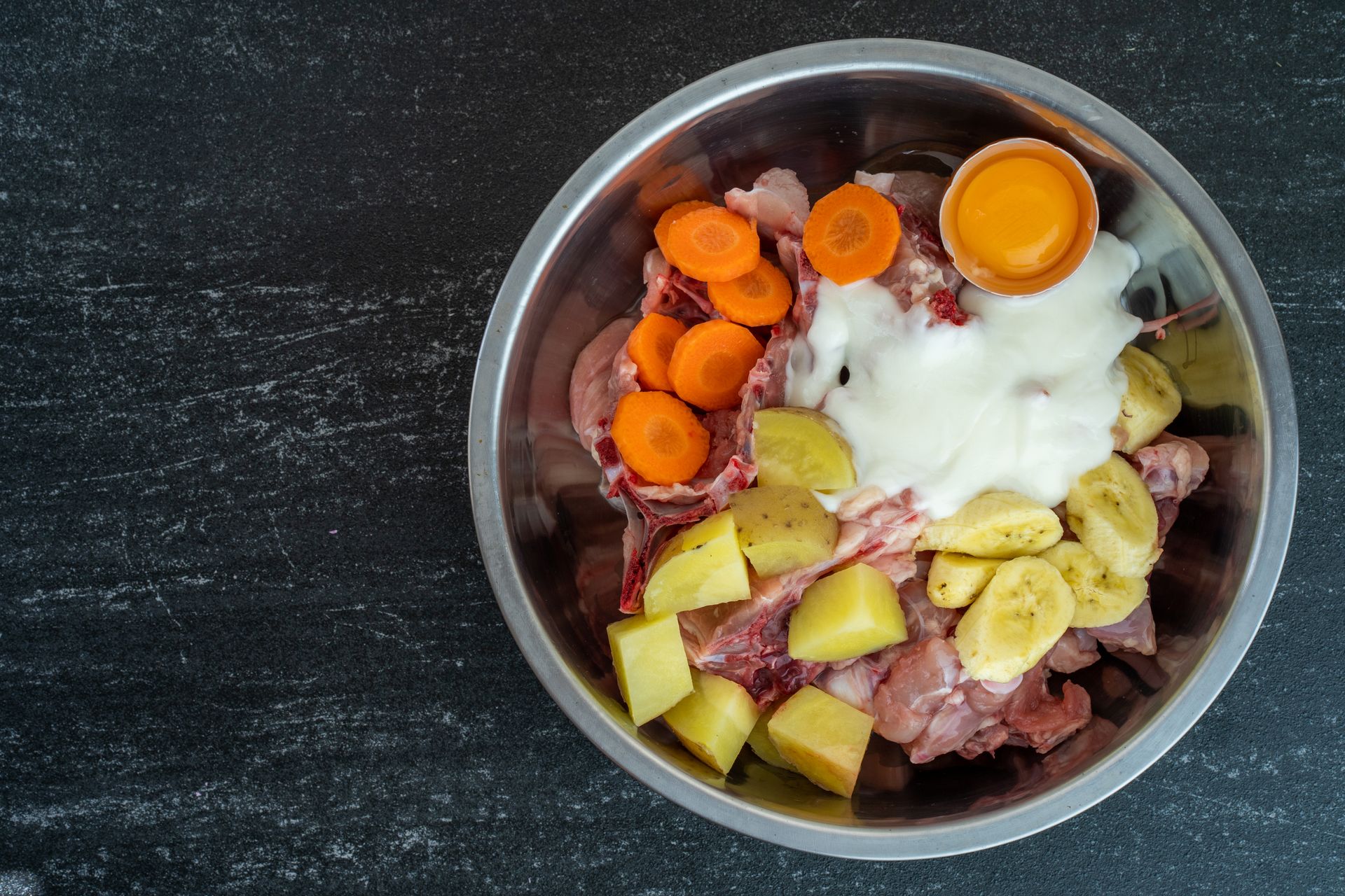 Stainless steel bowl with raw chicken, vegetables, and a cracked egg.