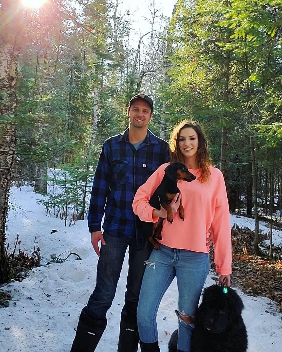 Couple posing with two dogs in a snowy forest. Man in blue plaid, woman holding dachshund, black dog at feet.