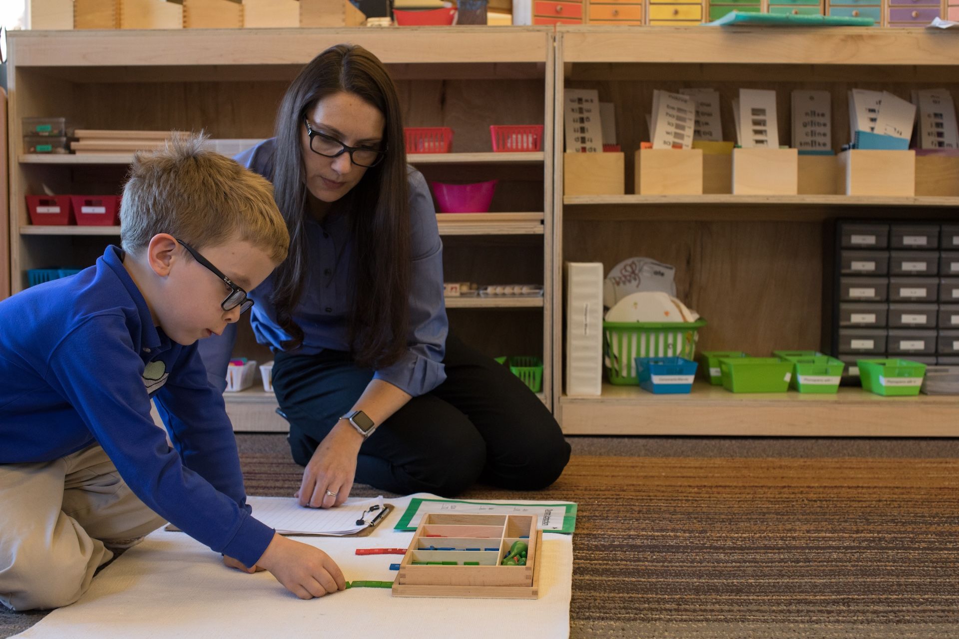 Montessori guide teaching a child, using educational materials on the floor. Both are focused and engaged, indoors.