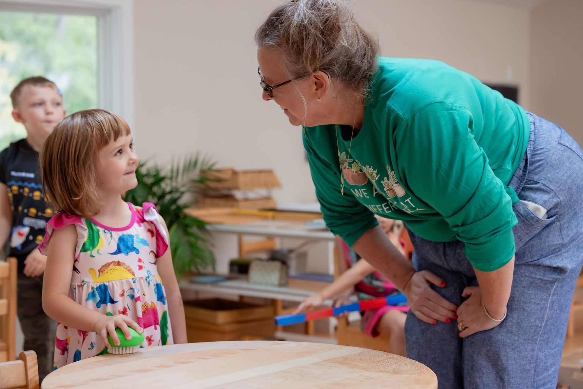 A Montessori guide leans to talk with a girl, indoors. Boy watches. Wooden table and shelves in background.