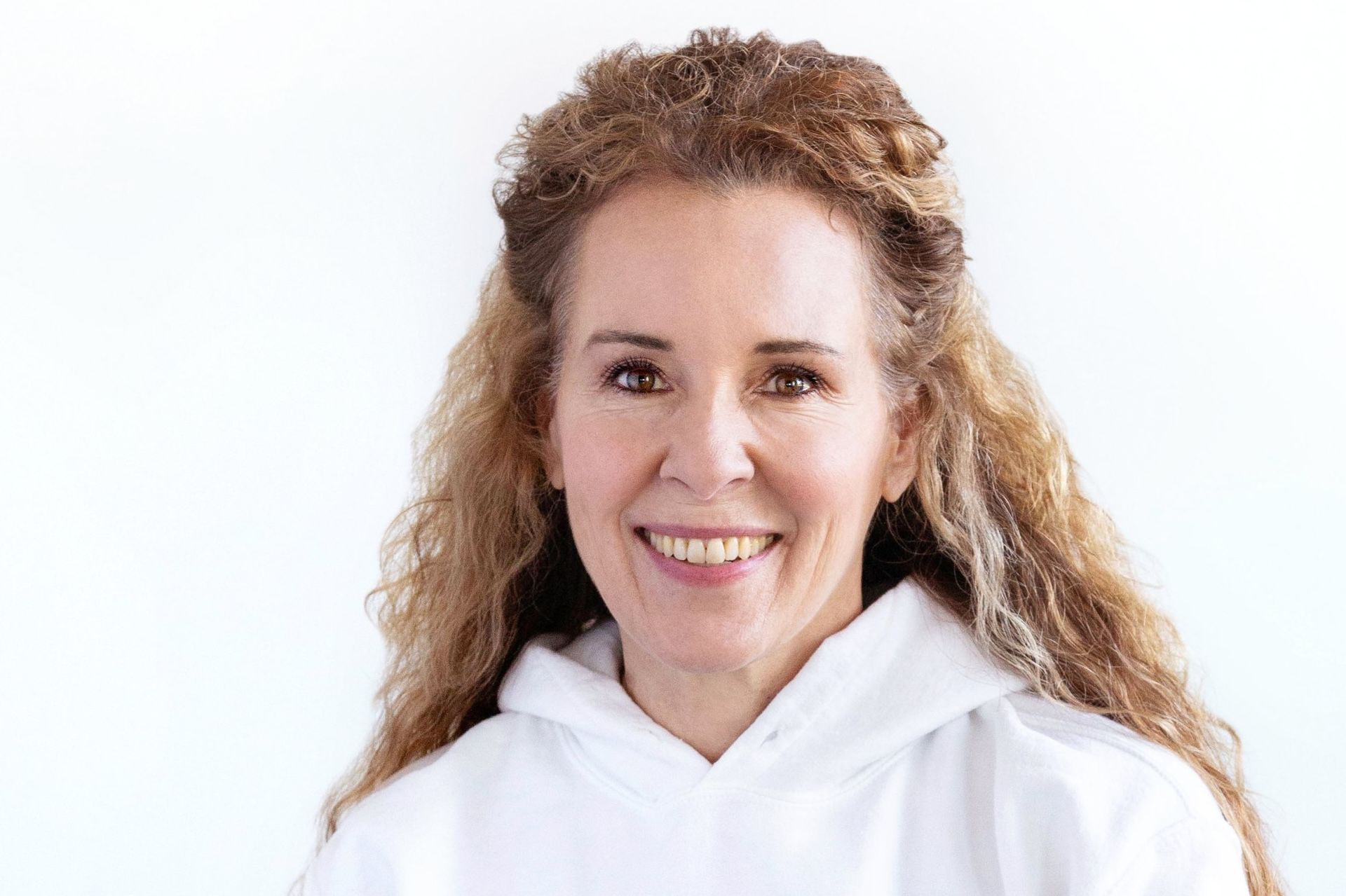 Woman with curly hair smiles, wearing a white hoodie, against a white backdrop.