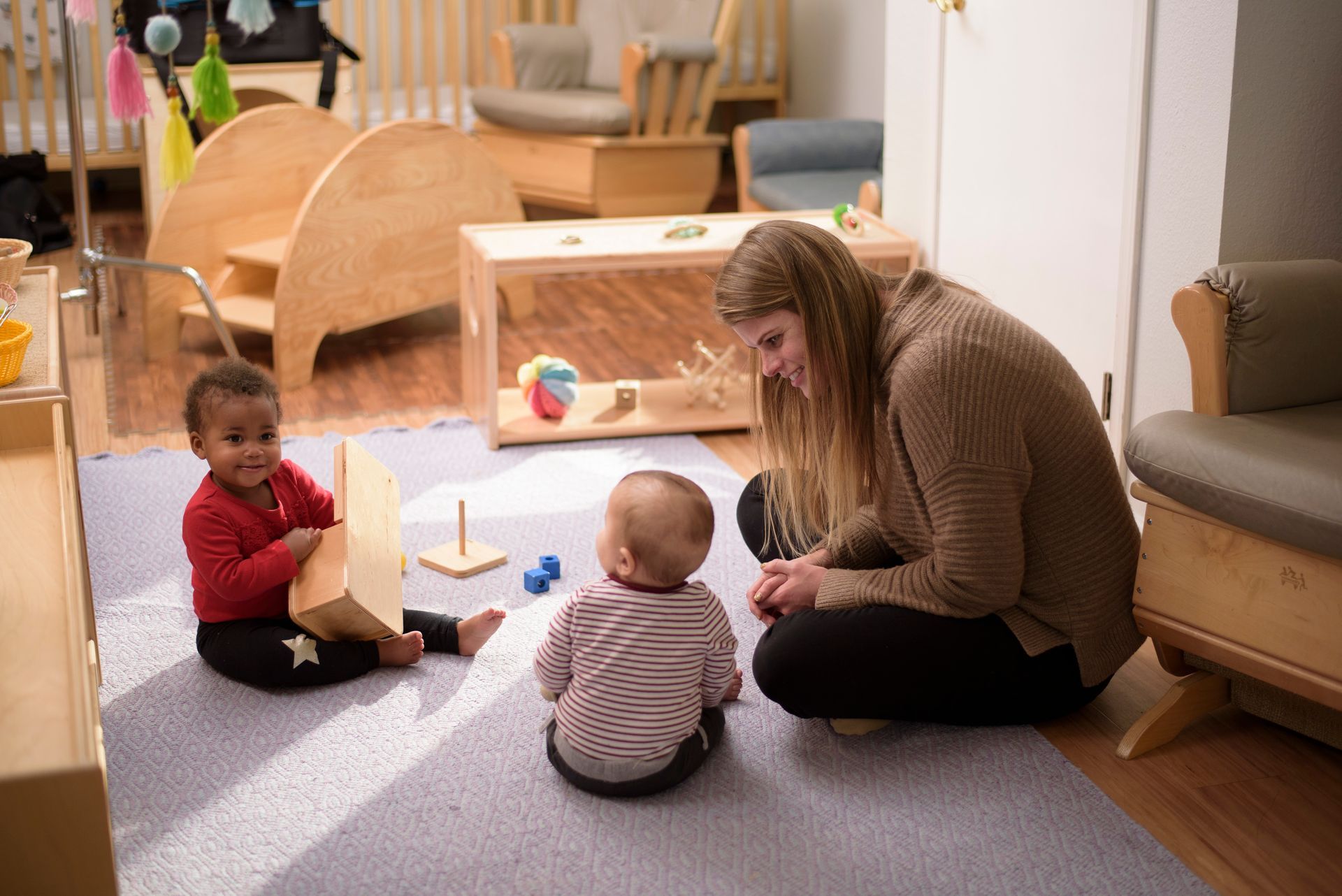 A Montessori guide and two Montessori children are playing on a blue rug in a brightly lit playroom.