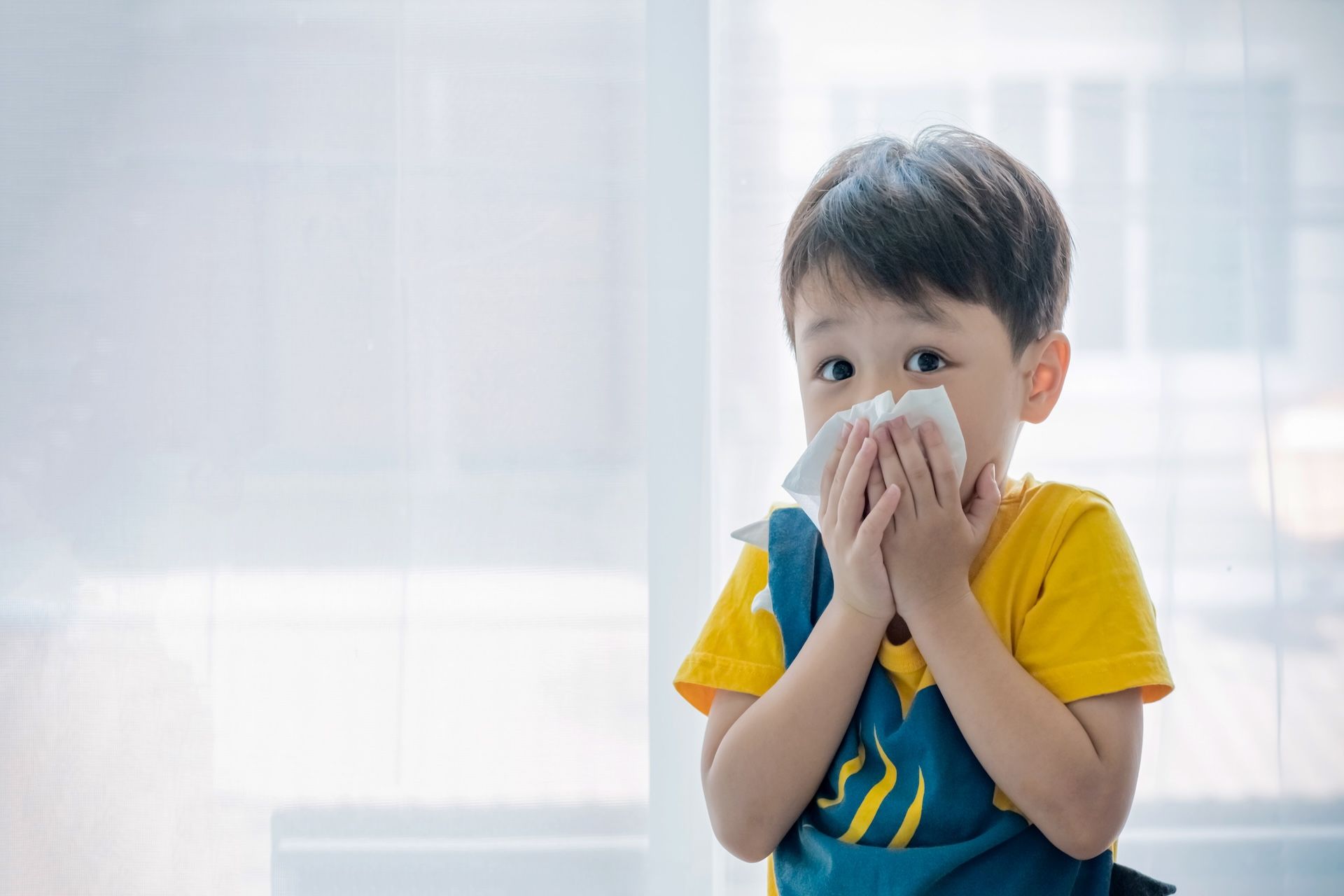 Montessori Child holding tissue to nose, sneezing. Wearing yellow shirt, blue overalls, indoors near a window.