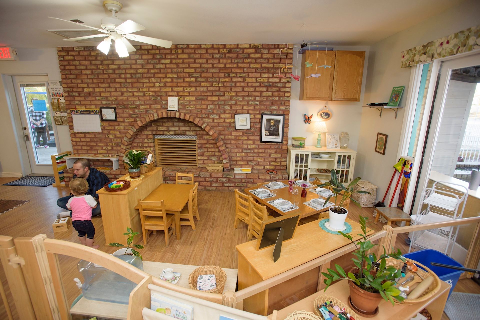 A brightly lit classroom with wooden furniture, brick wall, and a child and adult interacting.