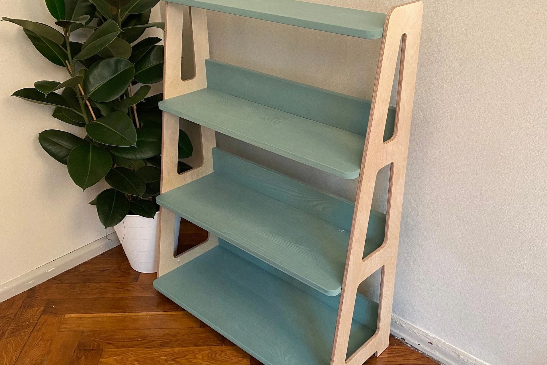 A teal and cream-colored wooden bookshelf next to a potted plant, sitting in a room.