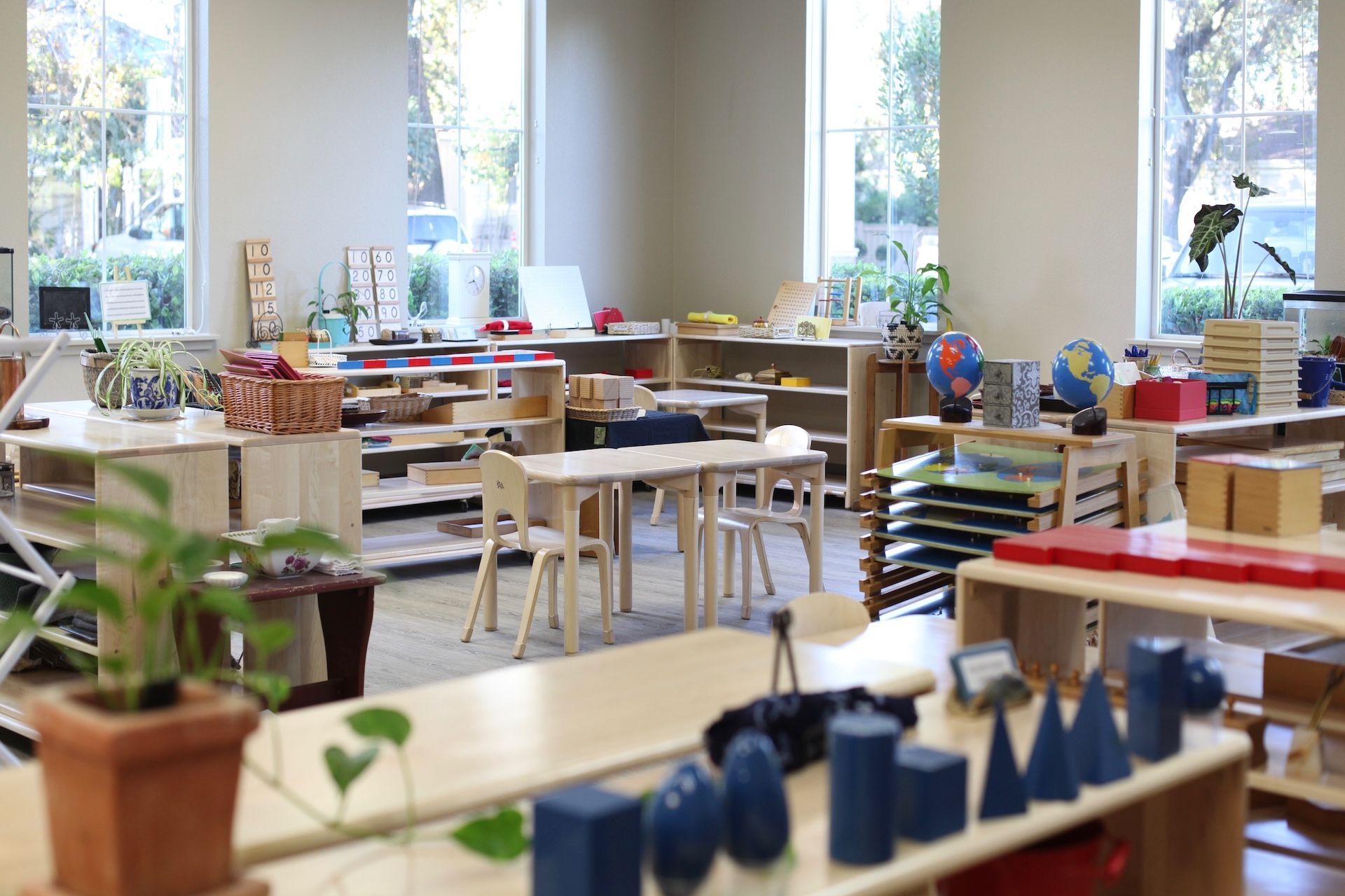Montessori classroom with wooden shelves, tables, and teaching materials near windows.