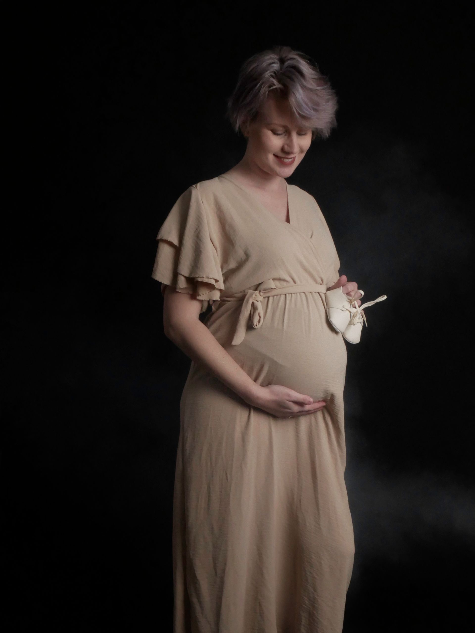 Een zwangere vrouw in een lange jurk houdt een paar babyschoentjes vast.
