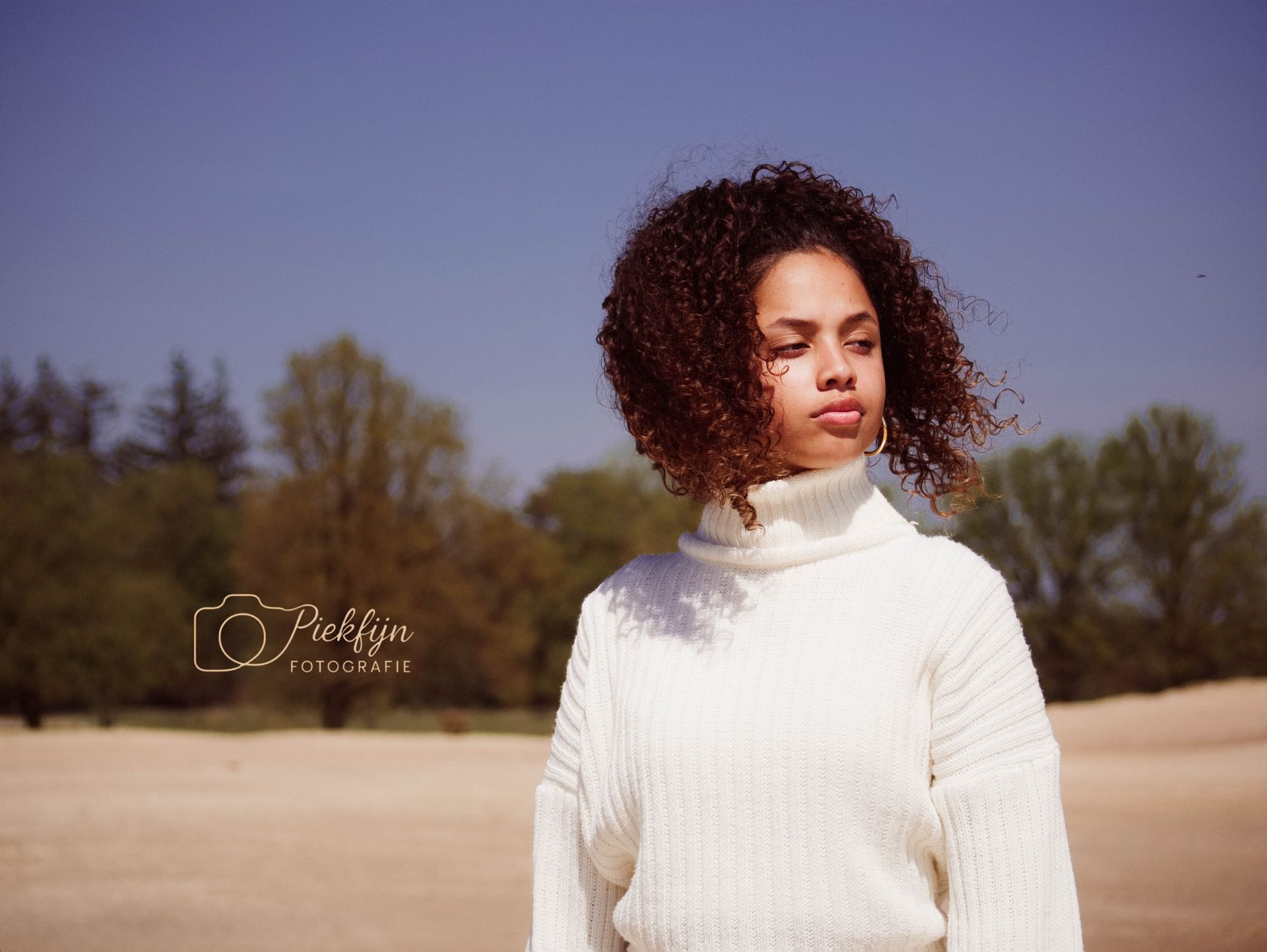 Een vrouw met krullend haar draagt ​​een witte trui en staat in een veld.