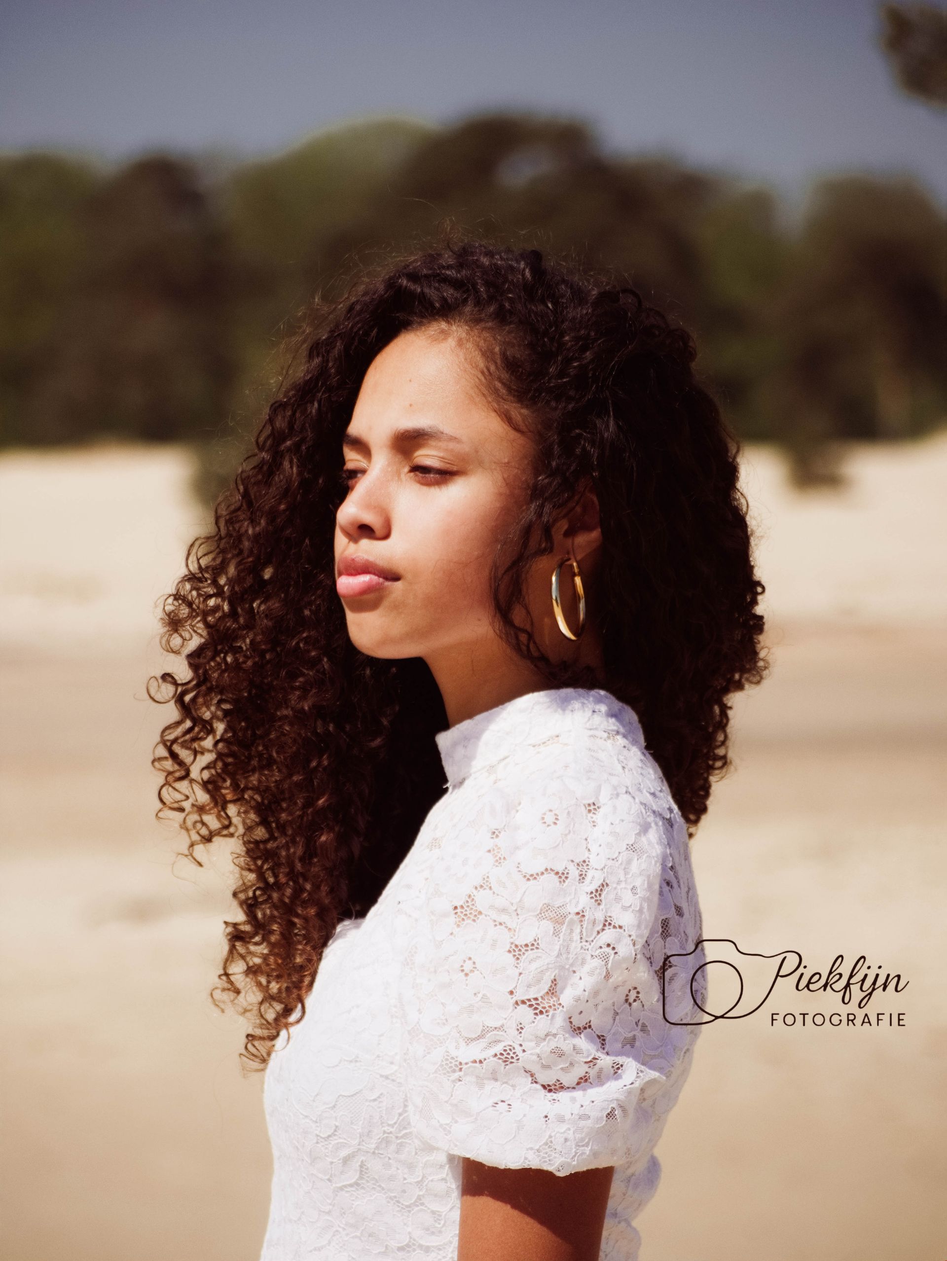 Een vrouw met krullend haar draagt ​​een wit kanten topje en gouden oorringen.