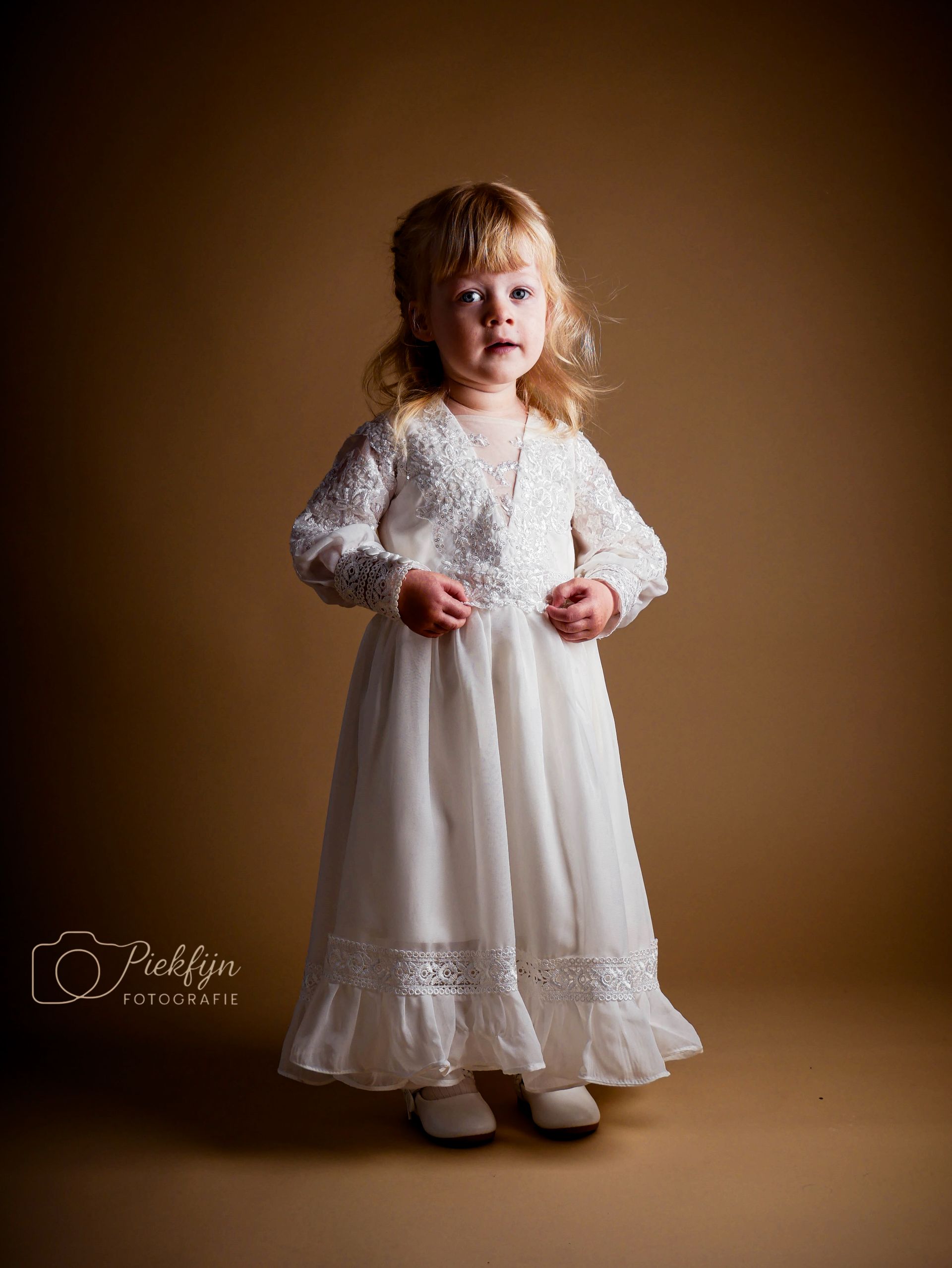 Een klein meisje in een witte jurk staat voor een bruine achtergrond.