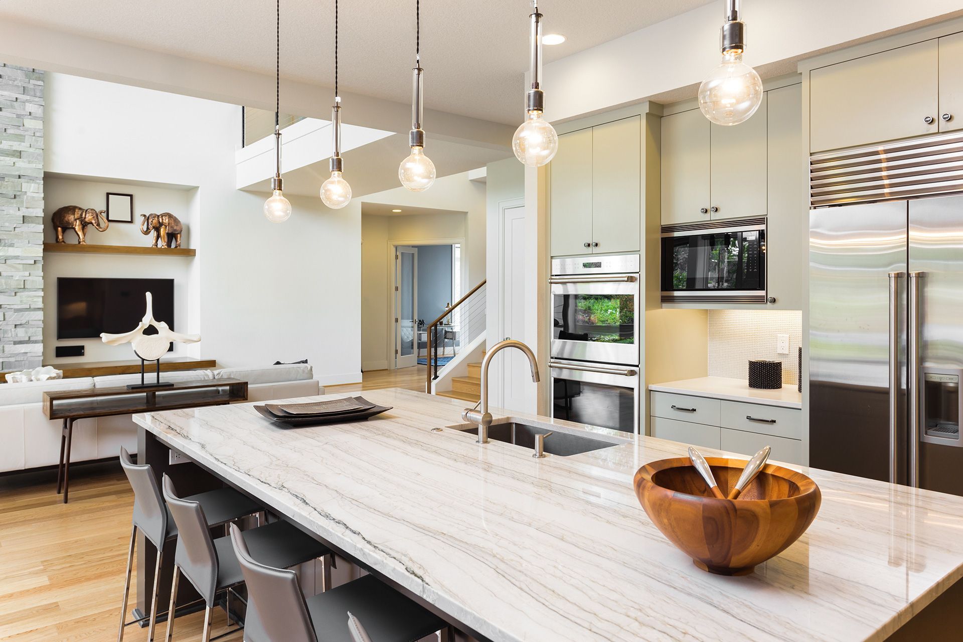 Modern open-plan kitchen with a large stone-topped island, wooden bowl, and pendant lighting, opening into a living room.