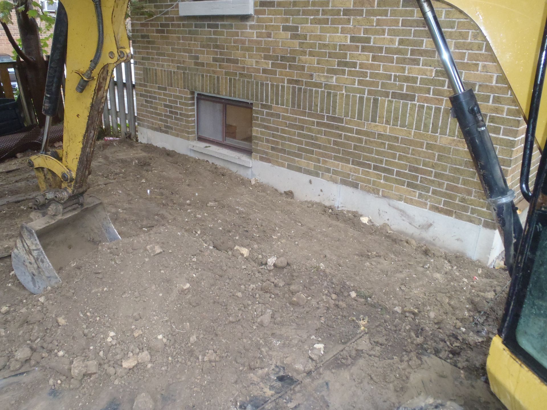 A yellow excavator is digging in front of a brick building