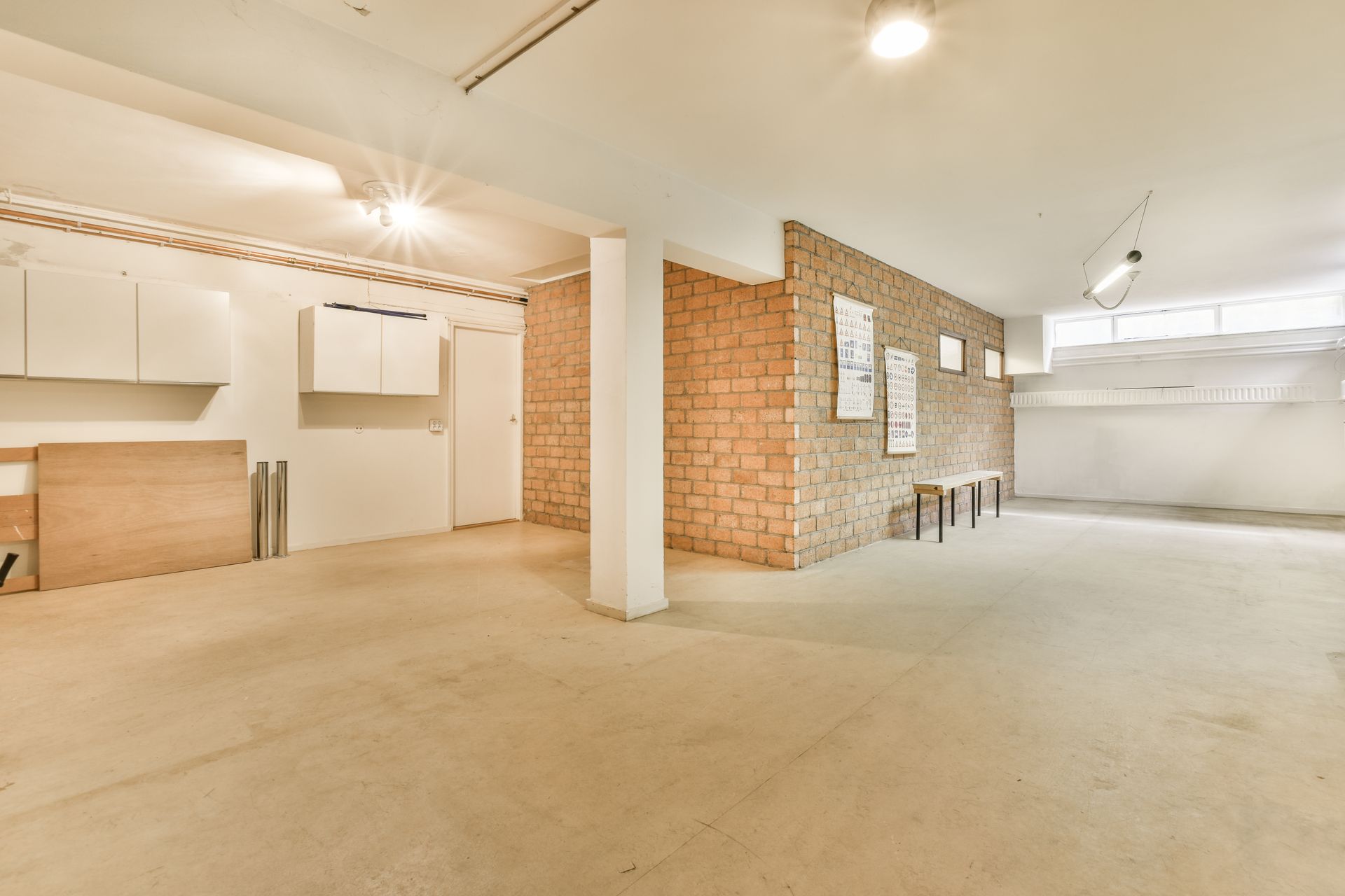 Empty, unfinished interior with brick wall, white support beam, cabinets, and concrete floor.