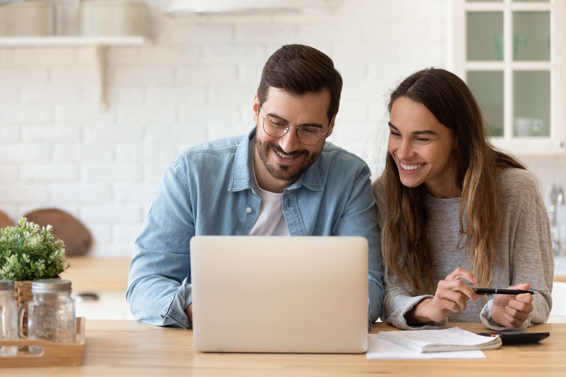 A couple smiles while looking at a laptop in a kitchen. The woman holds a pen over a notebook.