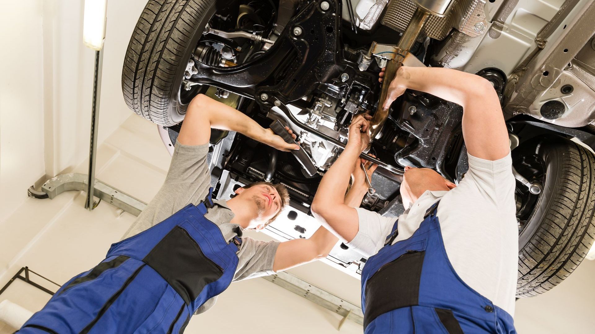 Two mechanics in blue coveralls work under a car in a shop.