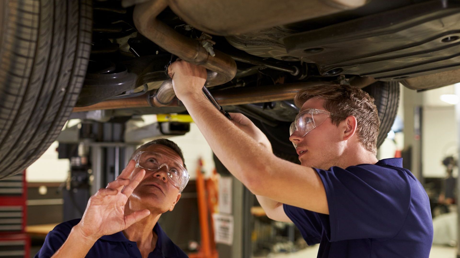 Two mechanics work under a car, examining exhaust system in a garage.