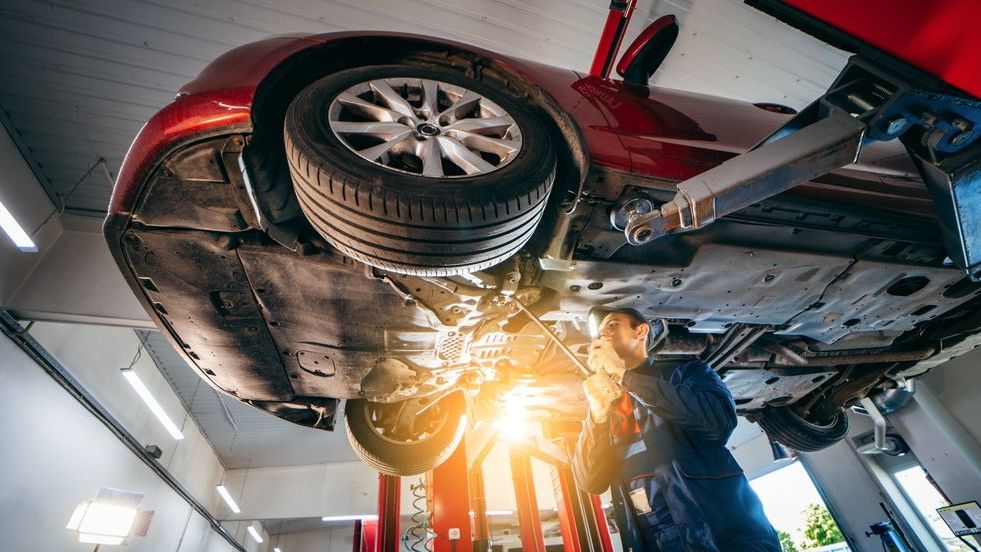 Mechanic working on a red car lifted on a hoist in a repair shop.