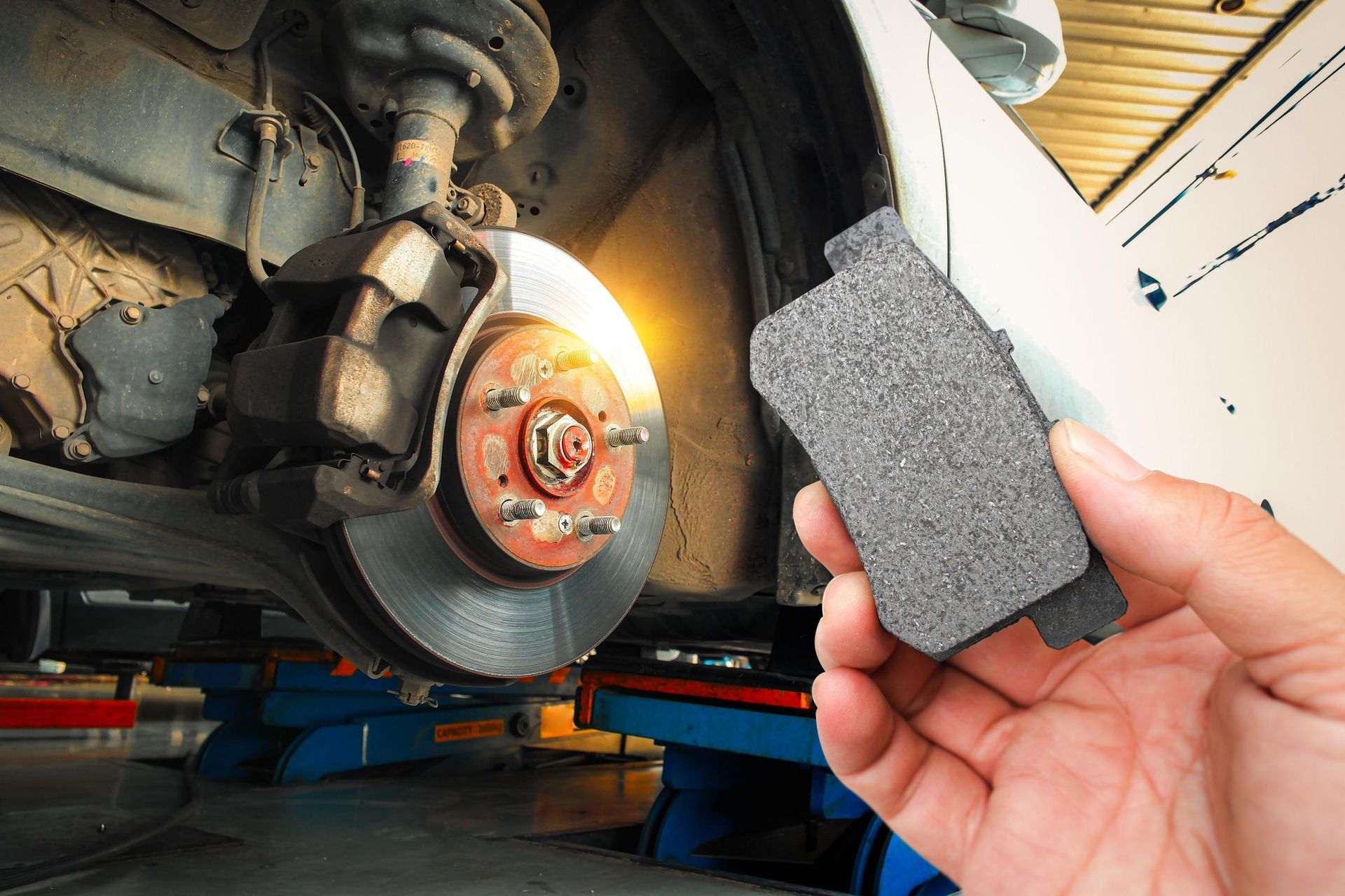 Hand holding a brake pad near a car’s exposed wheel brake assembly in a garage