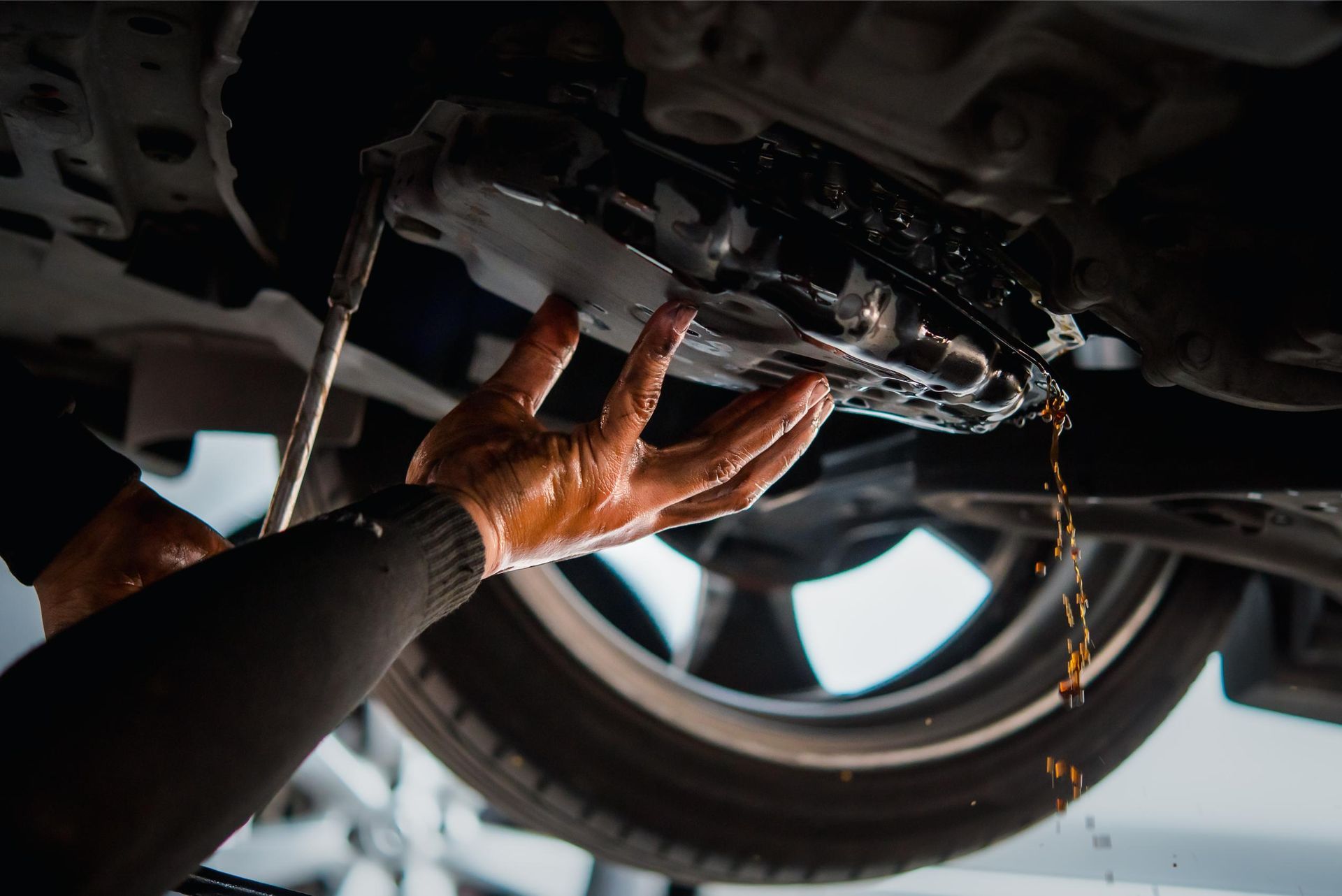 Mechanic draining oil from a car’s transmission pan under a vehicle. Oil dripping on hands; a tire visible.