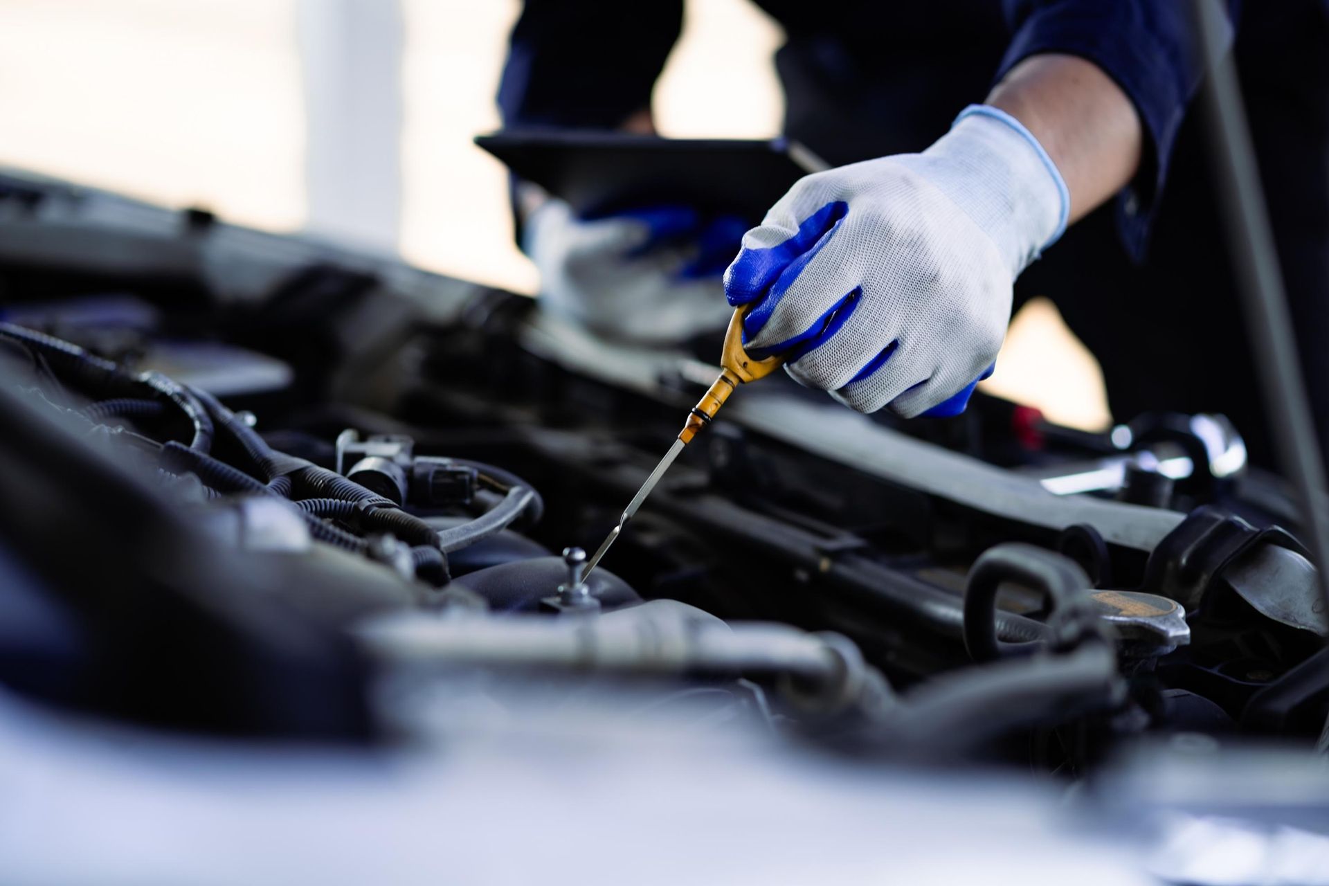 Mechanic checks oil dipstick in a car engine with gloved hand, holding a tablet.