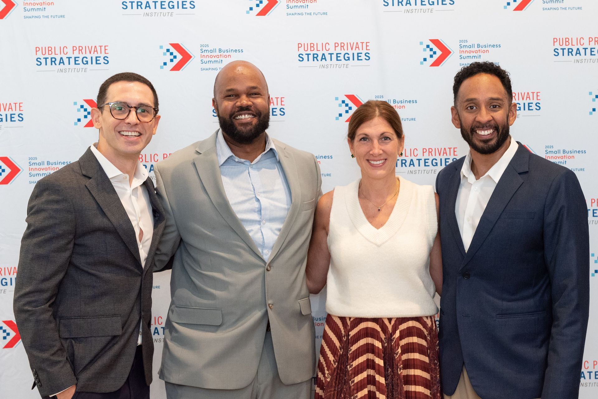 Four people pose together in front of a backdrop. Two men and two women smile. They are in business attire.