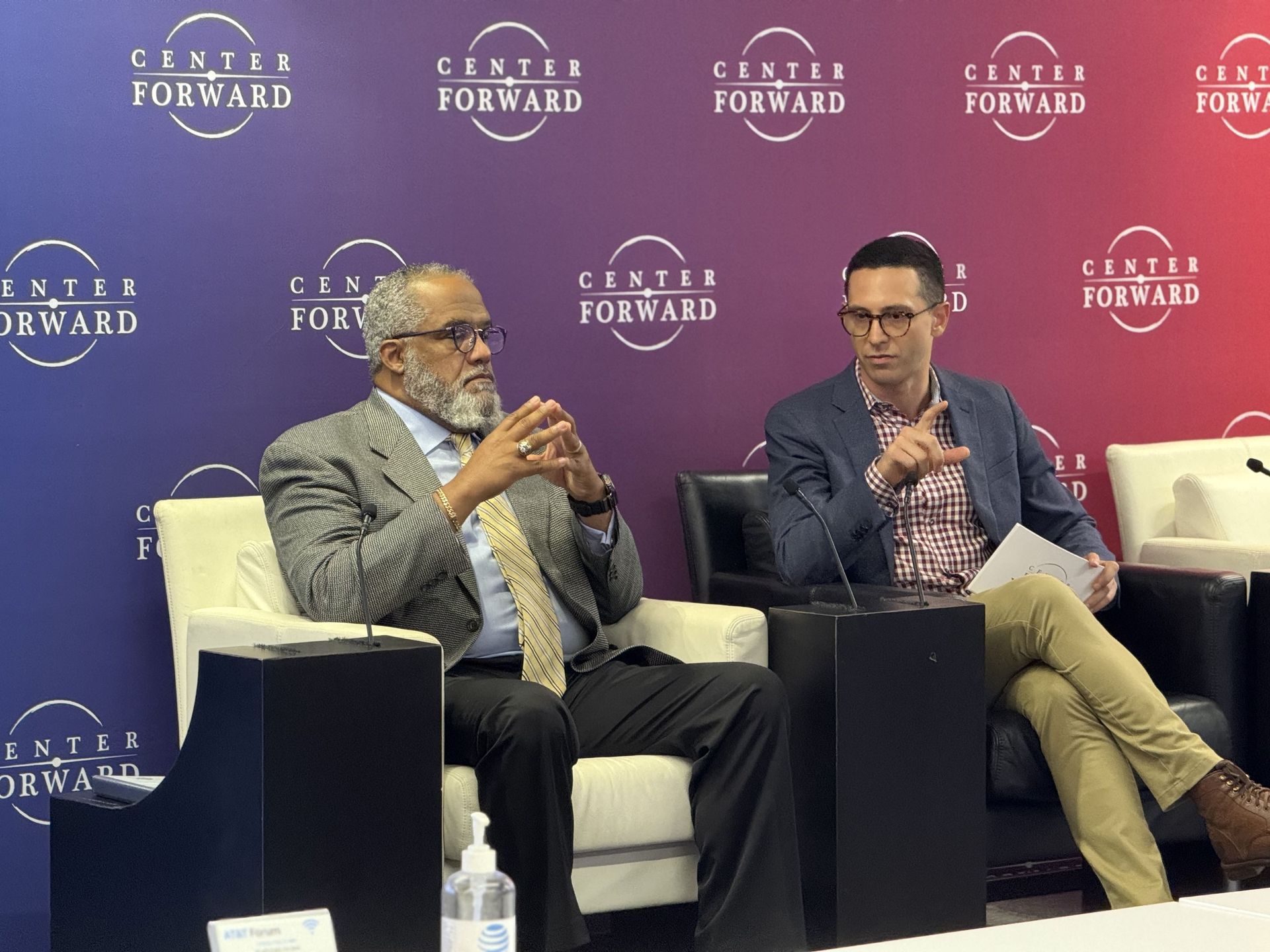 Two men in a discussion at a Center Forward event; seated on stage, microphone, blue and red background.