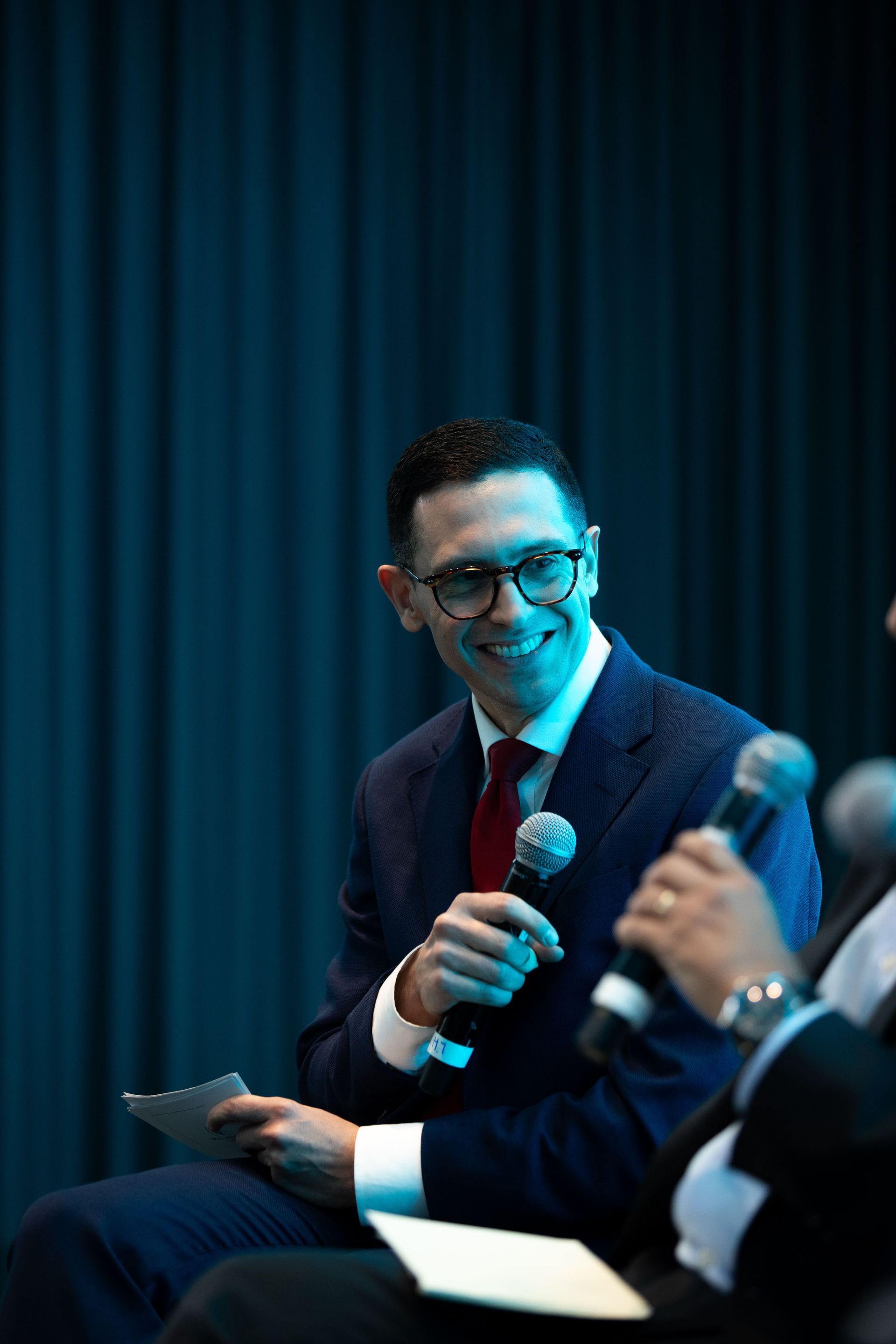 Man in suit with glasses smiles, holding microphone, looking at another person off-screen.