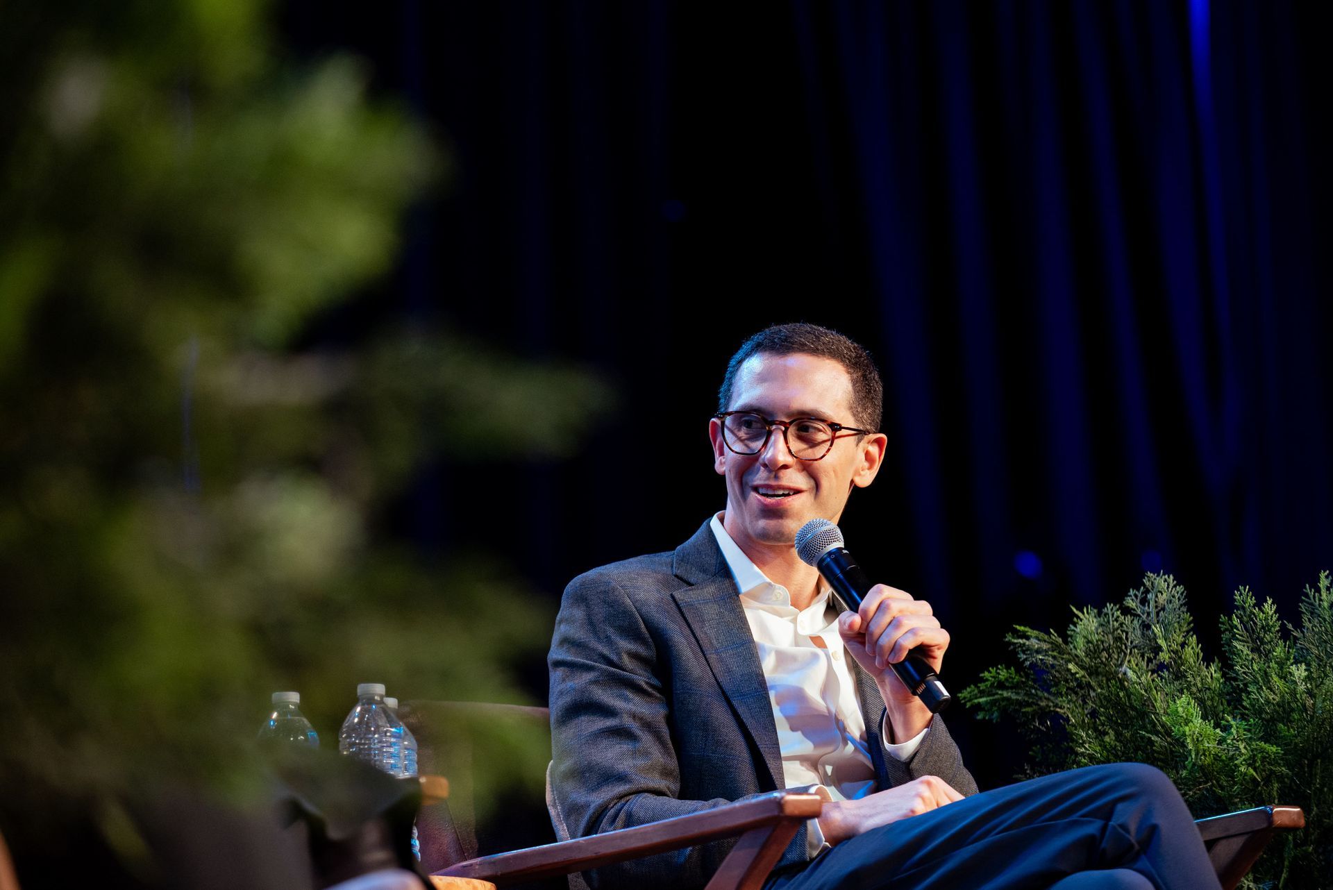 Man with glasses speaks into a microphone, sitting on stage with a dark background.