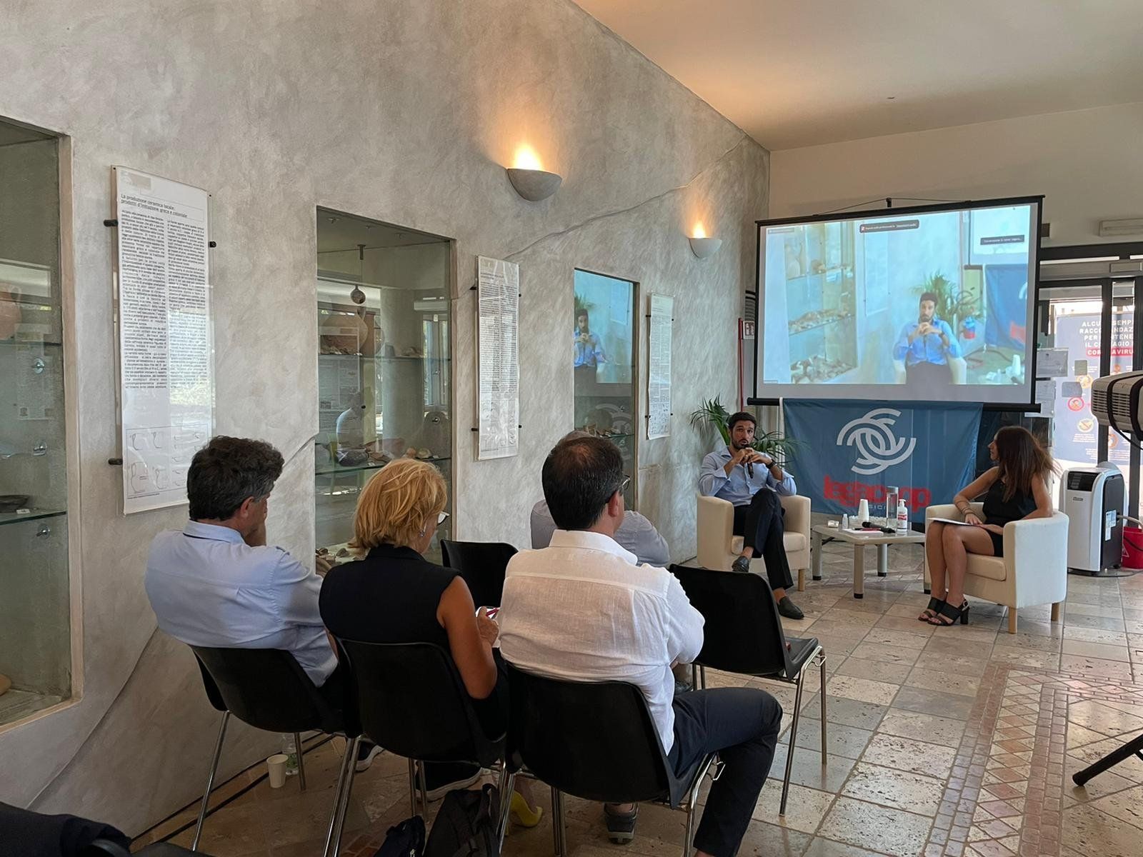 Un gruppo di persone è seduto su delle sedie davanti allo schermo di un proiettore.