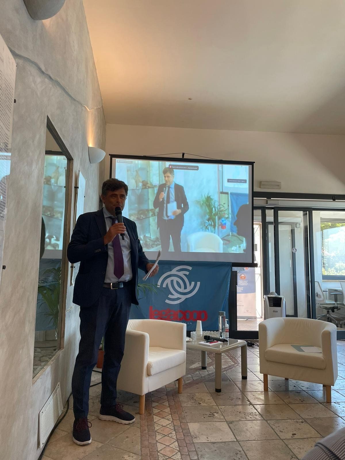 Un uomo è in piedi davanti allo schermo di un proiettore e parla in un microfono.